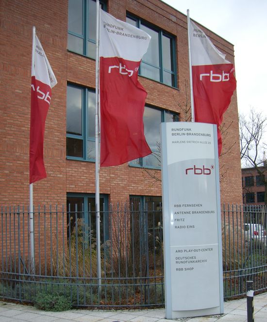Gezeigt wird ein Eingang des RBB-Studios in Potsdam mit drei RBB-Fahnen, die im Wind flattern und einem Hinweisschild am Eingang. – Zeichen auf Sturm? Fahnen flattern im Winde, wie die Aussagen des Senders gegenüber Gewerkschaften. Foto: Hirschler